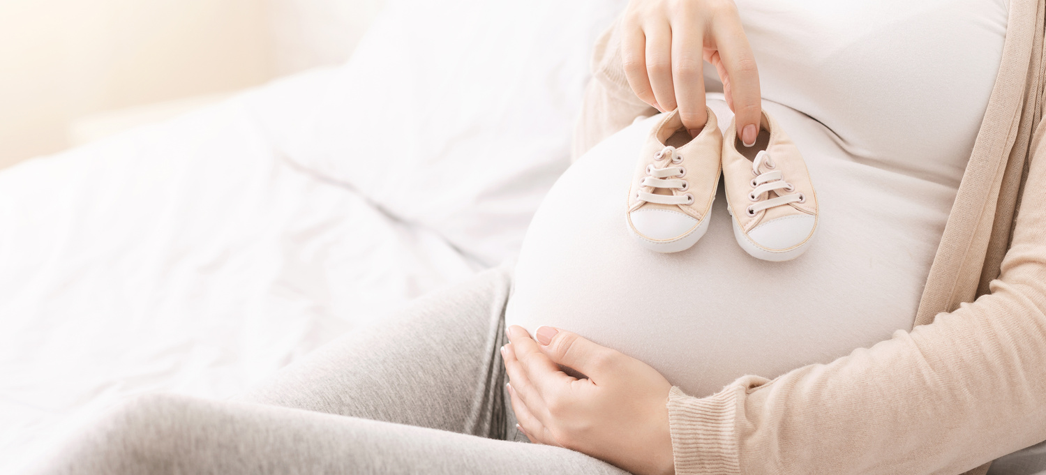 Бременна жена държи бебешки обувки върху корема си 