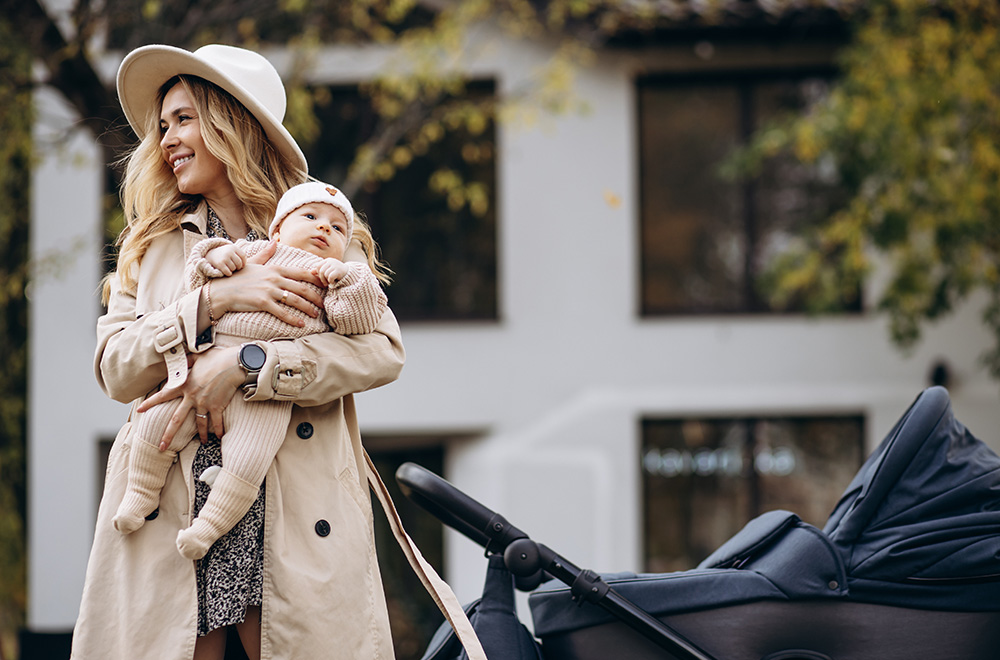 Жена държи бебе в ръцете си, докато върви до количка на фона на сграда.